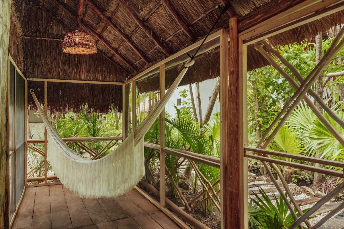 A hammock in a room with wooden floors and a thatched roof, overlooking a garden with various plants and a stone pathway.
