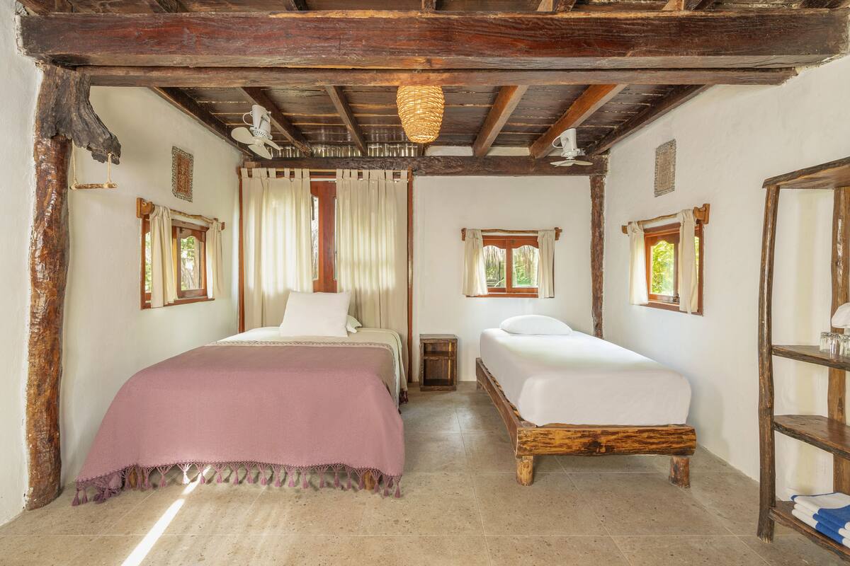 A rustic room with two beds, wooden beams, a hanging light fixture, and a small shelf with glasses.