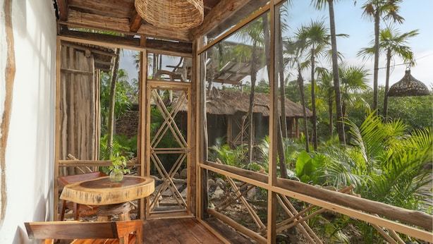 A wooden room with a round table and chairs, large windows, and a view of palm trees and thatched-roof huts. - Yaxchen Tulum Cabañas & Cenote (Tulum)