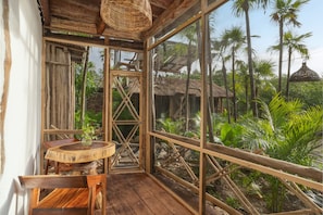 A wooden room with a round table and chairs, large windows, and a view of palm trees and thatched-roof huts.