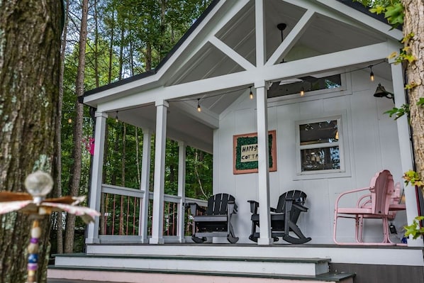 A white wooden structure with a porch, two black chairs, one pink chair, and a sign that reads 'HAPPY COTTAGE'.