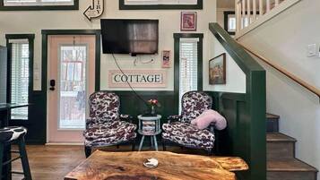 Un intérieur chaleureux avec une table basse en bois, deux fauteuils à motifs, une petite table d’appoint avec des livres et un escalier doté d’une rampe verte.