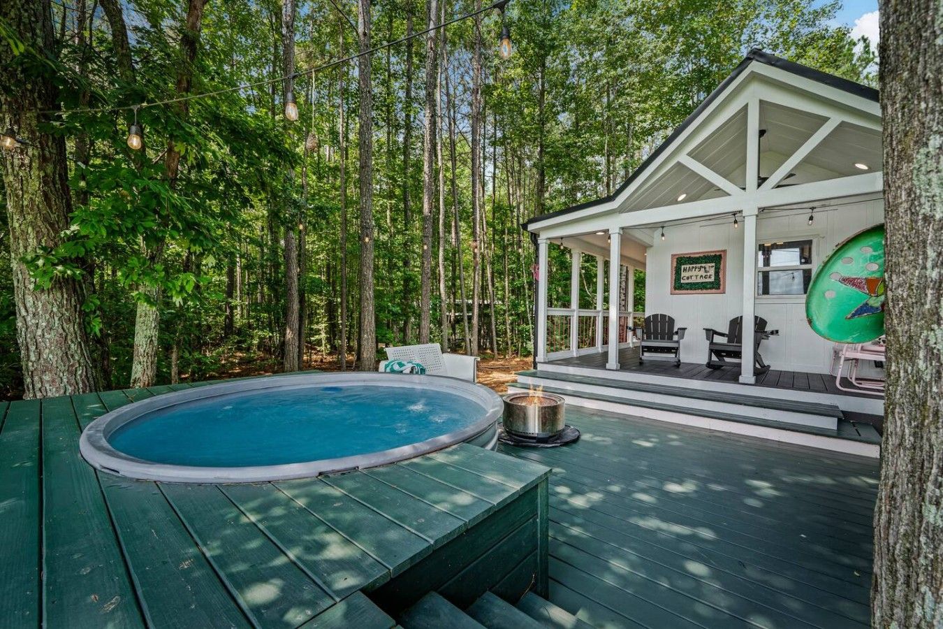 Une terrasse en bois avec un jacuzzi, entourée d’arbres, et un espace salon couvert avec une pancarte indiquant « HAPPY COTTAGE ».