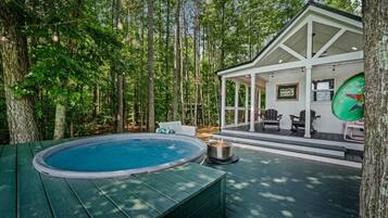 Une terrasse en bois avec un jacuzzi, entourée d’arbres, et un espace salon couvert avec une pancarte indiquant « HAPPY COTTAGE ».