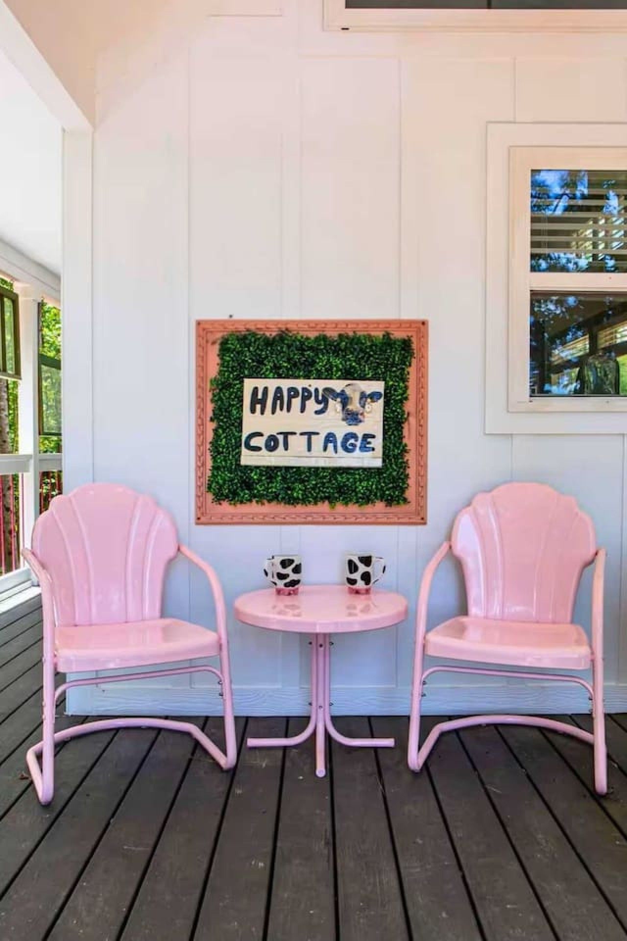 Un porche con dos sillas rosas y una mesita, un letrero que dice 'Happy Cottage', y una ventana con vista a follaje.