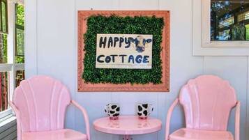 Une véranda avec deux chaises roses et une petite table, une enseigne indiquant «
Happy Cottage
», et une fenêtre donnant sur un paysage verdoyant.