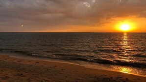 Ranta, jossa näkyy jalanjälkiä, jotka johtavat veteen, auringonlasku meren yllä ja pilvinen taivas.