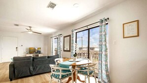 Un salon avec un canapé gris, une table à manger avec des chaises, un ventilateur de plafond et de grandes fenêtres ornées de rideaux à motifs.