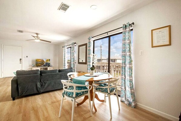 A living room with a grey sofa, a dining table with chairs, a ceiling fan, and large windows with patterned curtains.