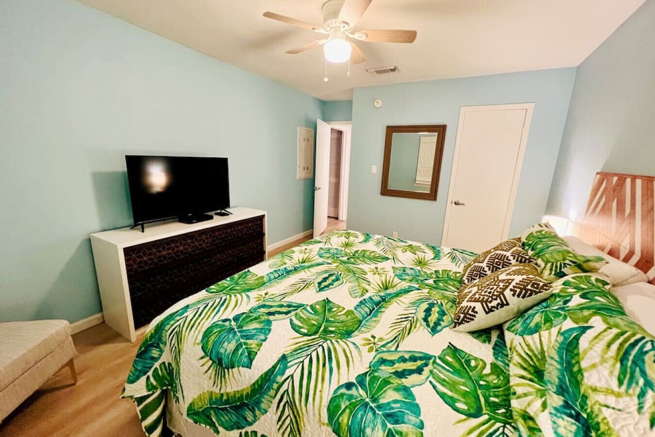 Ein Schlafzimmer mit Bett, Fernseher auf einer Kommode, Deckenventilator und einer Tür.