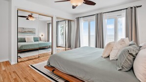 Ein Schlafzimmer mit Bett, Deckenventilator, großem Fenster mit Vorhängen und Blick ins Freie.