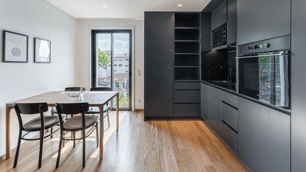 Une cuisine moderne avec des meubles de couleur sombre, un four encastré et un coin repas comprenant une table et des chaises en bois.