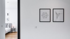 Un couloir orné de deux œuvres d’art géométriques murales, un lit avec une tête de lit grise, une table de chevet avec une lampe et un plancher en bois.