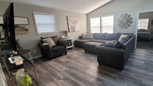 A modern living room with a sectional sofa, a coffee table, and a TV. There are two windows with blinds, a decorative wall piece, and a door leading to another room. - Harmony House (Kokomo)