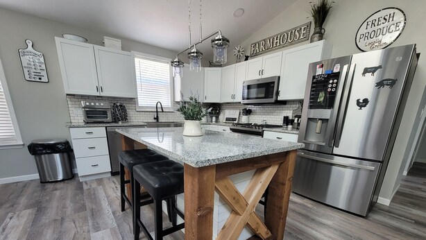 A modern kitchen with white cabinets, stainless steel appliances, a wooden island with bar stools, and a sign reading "FARMHOUSE". - Harmony House (Kokomo)