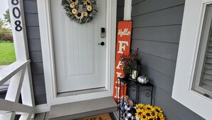 A front door with a wreath, a 'Hello' sign, a doormat, and a planter with sunflowers. - Harmony House (Kokomo)