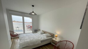 A modern bedroom with a large bed, a bedside table with a lamp, a chair, and a view of the outdoors through a sliding door. - New appartment near the center for 6 people with car park (LACANAU OCEAN)