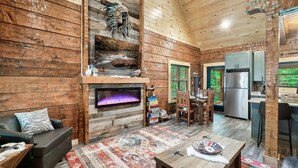 A log cabin interior with a fireplace, dining area, kitchen, and a large window with a view of greenery. - Rustic 2BR Log Cabin w/ Private Indoor Pool & Hot Tub — Near Pigeon Forge (Sevierville)