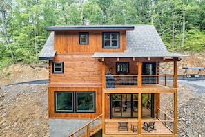 Une cabane en rondins à deux étages, dotée d’un balcon et d’une terrasse, entourée de forêt.