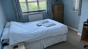A neatly made bed with white linens, a bedside table with a remote control, a dresser, and a window with curtains. - Captivating Apartment Near Gatwick (Crawley)