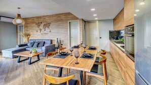 A modern living space with a wooden accent wall, a dining area with a wooden table and chairs, and a kitchen with stainless steel appliances. - 2-bed Pool Villa in Panormo (Mylopotamos)