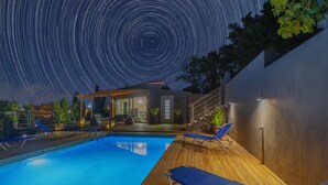 A swimming pool with lounge chairs, a wooden deck, and a building with a glass door. - 2-bed Pool Villa in Panormo (Mylopotamos)
