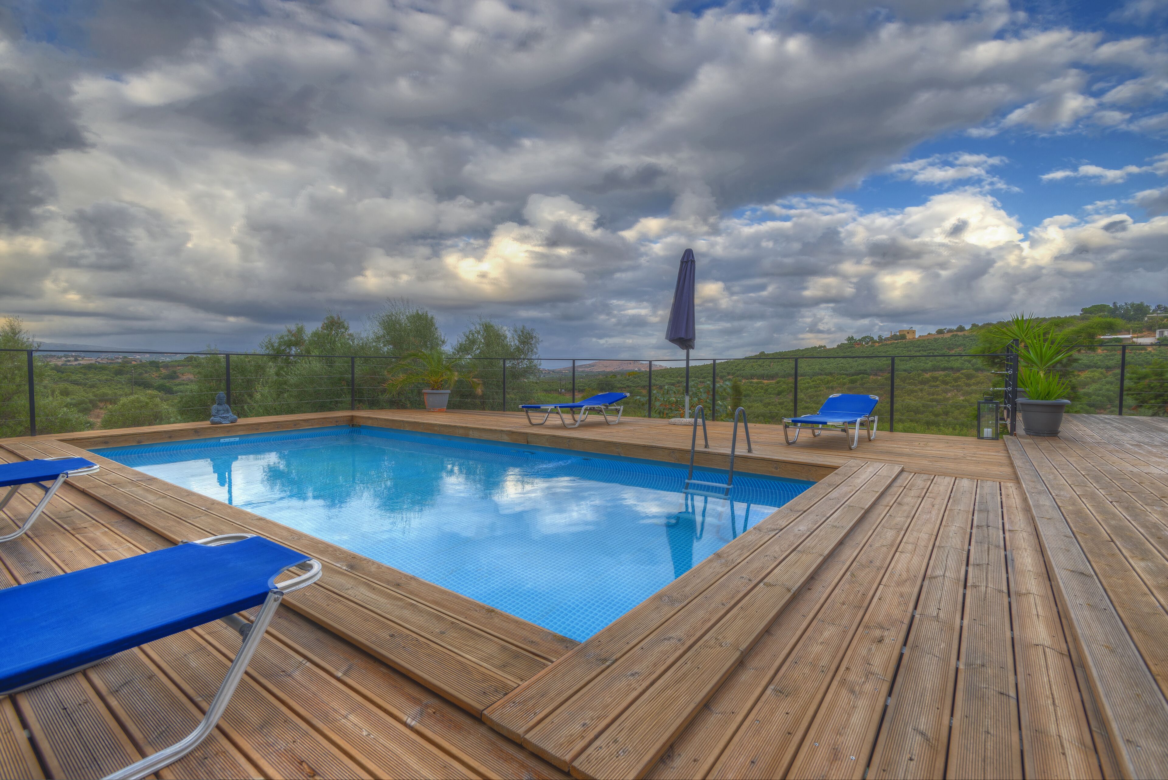 A wooden deck with a swimming pool, blue lounge chairs, and a view of the landscape.