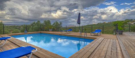 A wooden deck with a swimming pool, blue lounge chairs, and a view of the landscape.