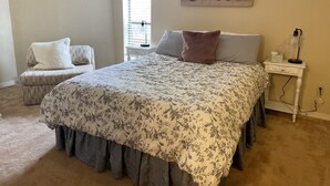 Ein Schlafzimmer mit einem Bett, einem Sessel, einem Nachttisch, einer Lampe und einem Fenster.