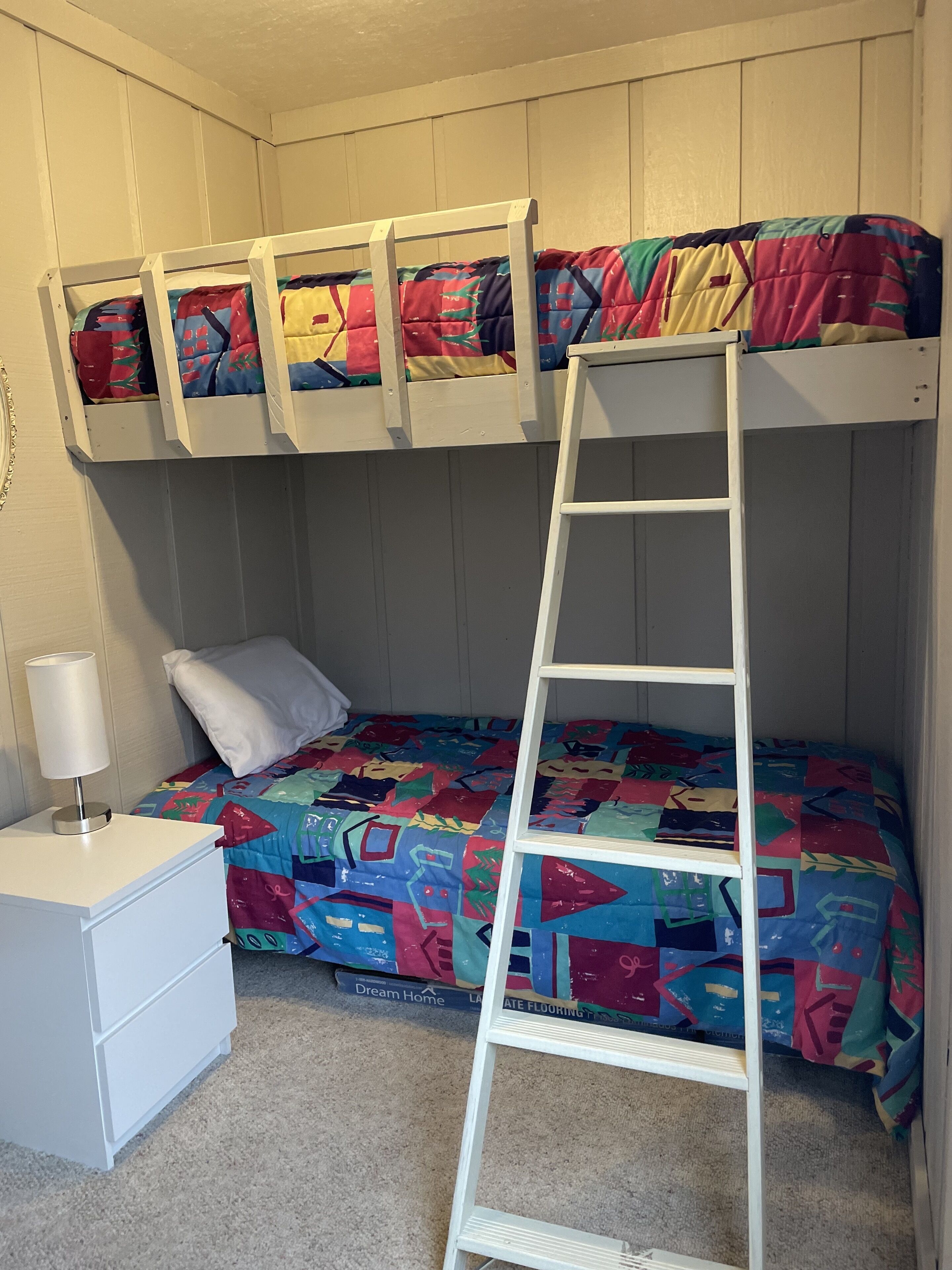 Un lit superposé avec une échelle, une table de chevet avec une lampe et une couverture colorée.
