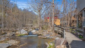 A wooden bridge over a clear stream, surrounded by trees and a building with a red roof. - Still Standing! Cozy Cottage, Incredible Location! (Lake Lure)
