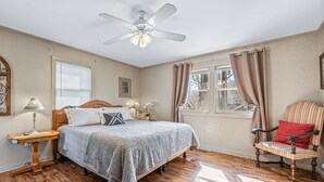 A bedroom with a wooden floor, a bed with a white and gray blanket, a nightstand with a lamp, a chair with a red pillow, a ceiling fan, and a window with curtains. - Still Standing! Cozy Cottage, Incredible Location! (Lake Lure)