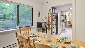 Un comedor con una mesa de madera dispuesta para cuatro personas, un florero con ramas y vistas al bosque por la ventana.