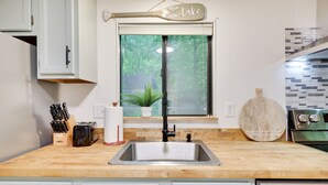 Una cocina con encimera de madera, pileta de acero inoxidable, ventana con vista a árboles y un cartel sobre la ventana que dice 'Llévame al lago'.