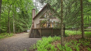Una cabaña con terraza de madera y chimenea, rodeada de un bosque frondoso.