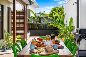 An outdoor dining area with a wooden table, green chairs, and a variety of food and drinks. - Little Fern House a Tropical Beach Hideaway Mudjimba,  Sunshine Coast 🛫 5 mins (Mudjimba)