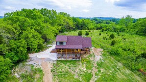 A two-story house with a large porch and a covered outdoor area, surrounded by lush greenery and hills in the background. - Gorgeous custom log cabin on 185 acres (Dellrose)