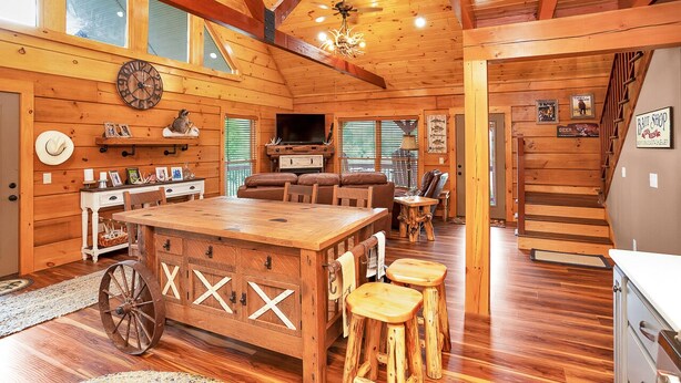 A rustic cabin interior with a wooden dining table, bar stools, a fireplace, and a loft bedroom with a staircase. - Gorgeous custom log cabin on 185 acres (Dellrose)