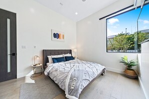 Une chambre moderne avec un grand lit, des tables de chevet, une œuvre d’art accrochée au mur, une fenêtre donnant sur un beau paysage et une plante en pot.