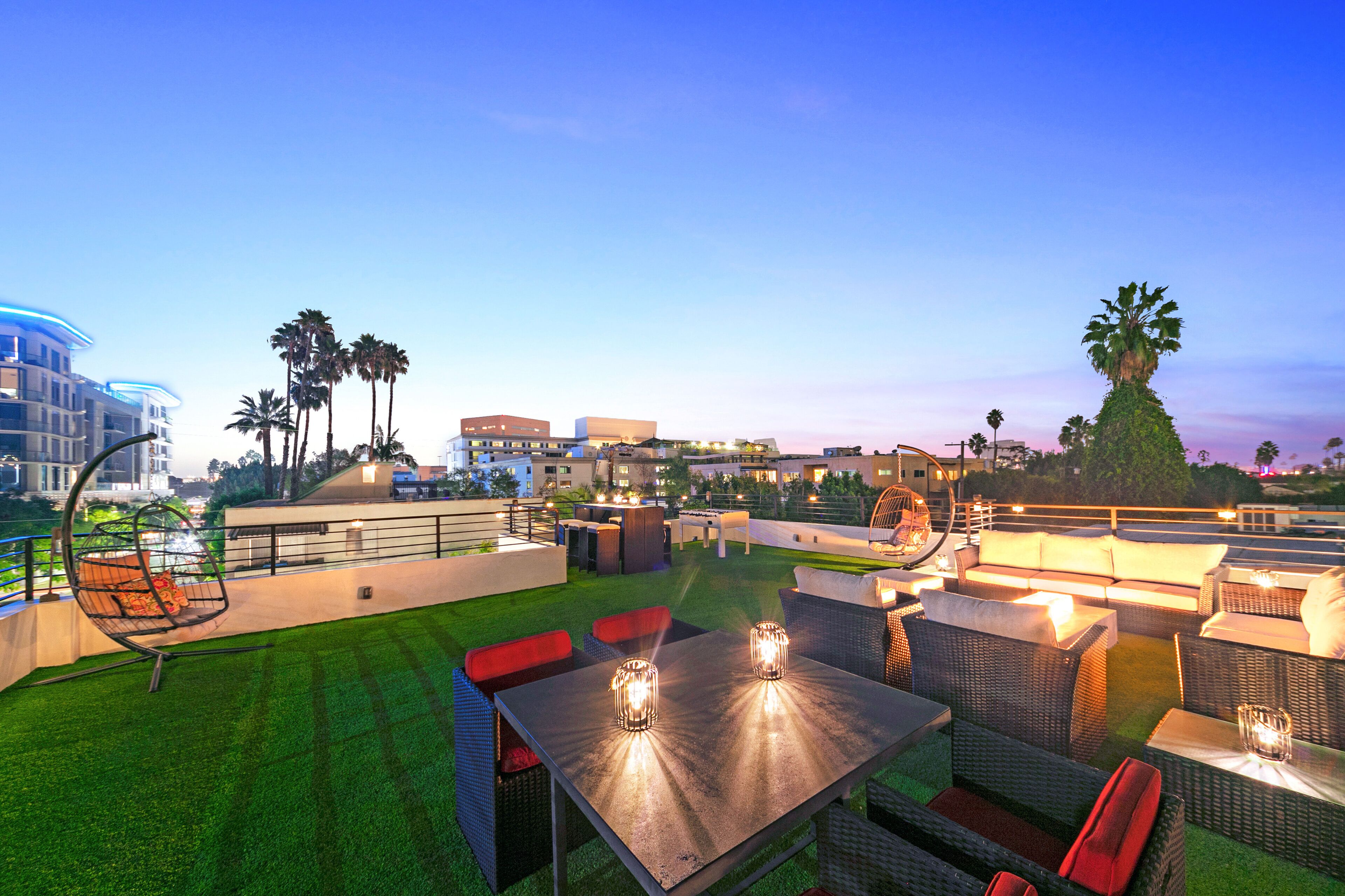An outdoor patio area with seating and tables, surrounded by palm trees and buildings in the background.