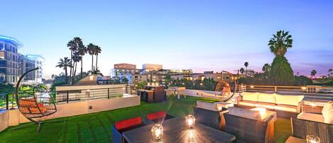 An outdoor patio area with seating and tables, surrounded by palm trees and buildings in the background.