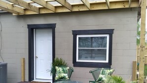 A covered patio with a wooden pergola, a door, a window, two green chairs, a small table, and potted plants. - Restored Building in Downtown Lincoln- Your Gateway to the Lake! (Lincoln)