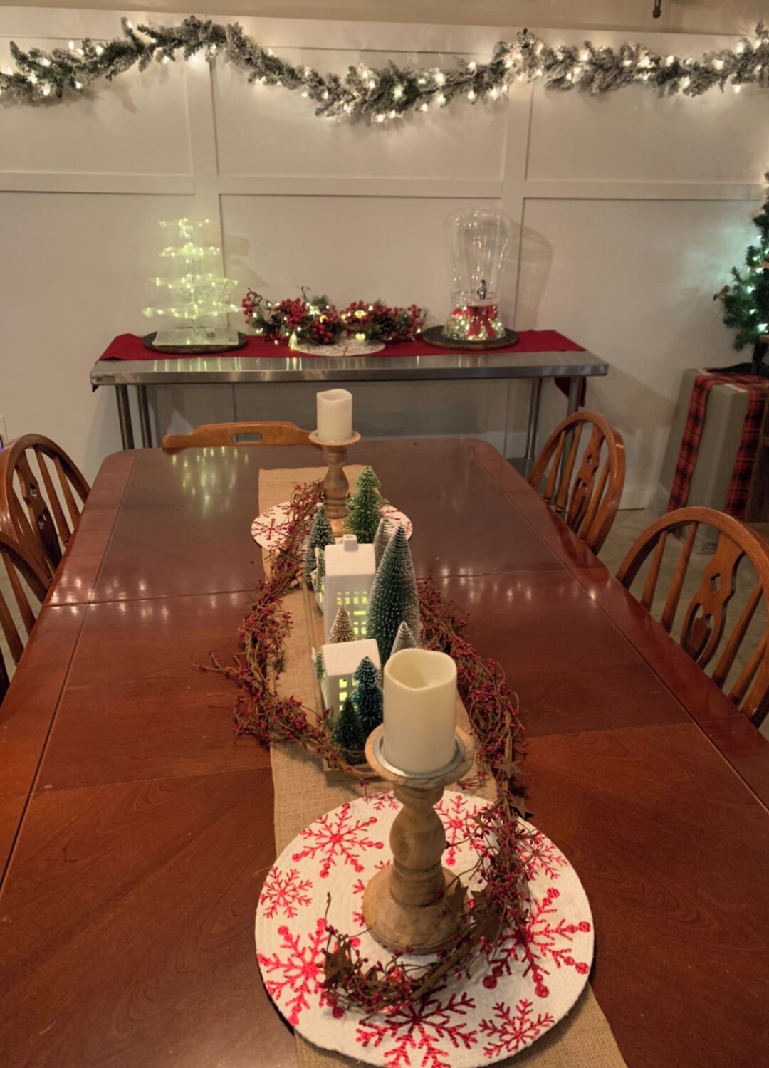 A dining table with a centerpiece featuring candles, miniature Christmas trees, and a garland.