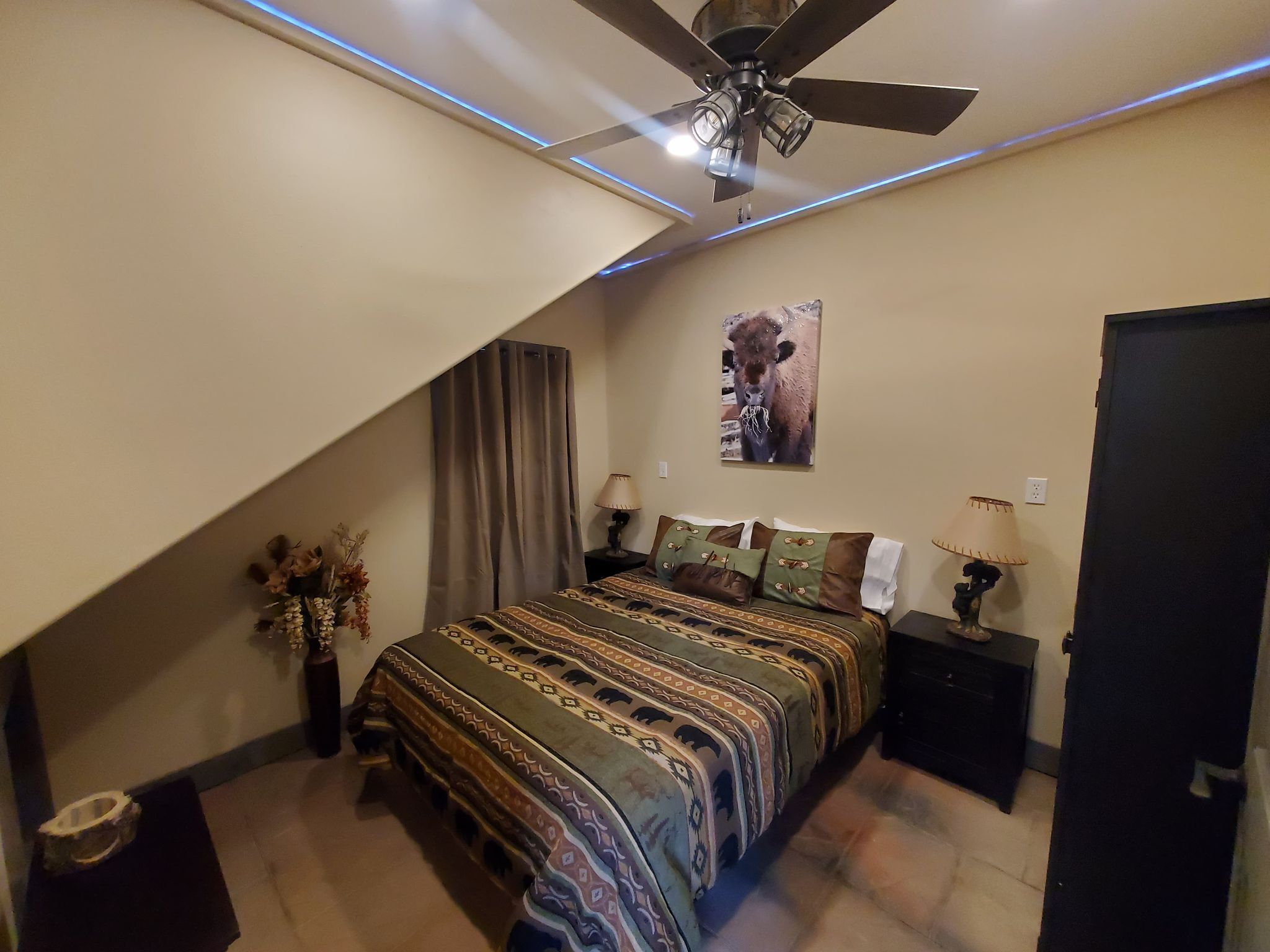 A bedroom with a bed, a ceiling fan, a wall-mounted lamp, and a painting.