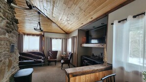 A cozy room with a stone wall, wooden ceiling, a fireplace, a sofa, a small table, and a bar area. - Remodeled Historic Cabin (West Yellowstone)