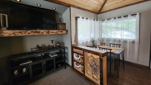 A cozy cabin interior with a wooden ceiling, a large flat-screen TV mounted on a wall, a dining area with a wooden table and chairs, and a small kitchen area with a built-in refrigerator and shelves. - Remodeled Historic Cabin (West Yellowstone)