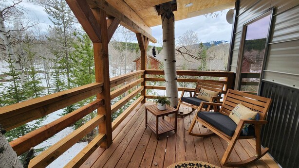 A wooden deck with a wooden bench and a rocking chair, a small table with a plant, and a view of trees and a body of water. - Remodeled Historic Cabin (West Yellowstone)