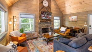 A cozy cabin interior with a stone fireplace, wooden walls, a dining area, and a large window.