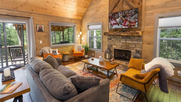 A cozy cabin interior with a stone fireplace, wooden walls, and a large window overlooking a forest.