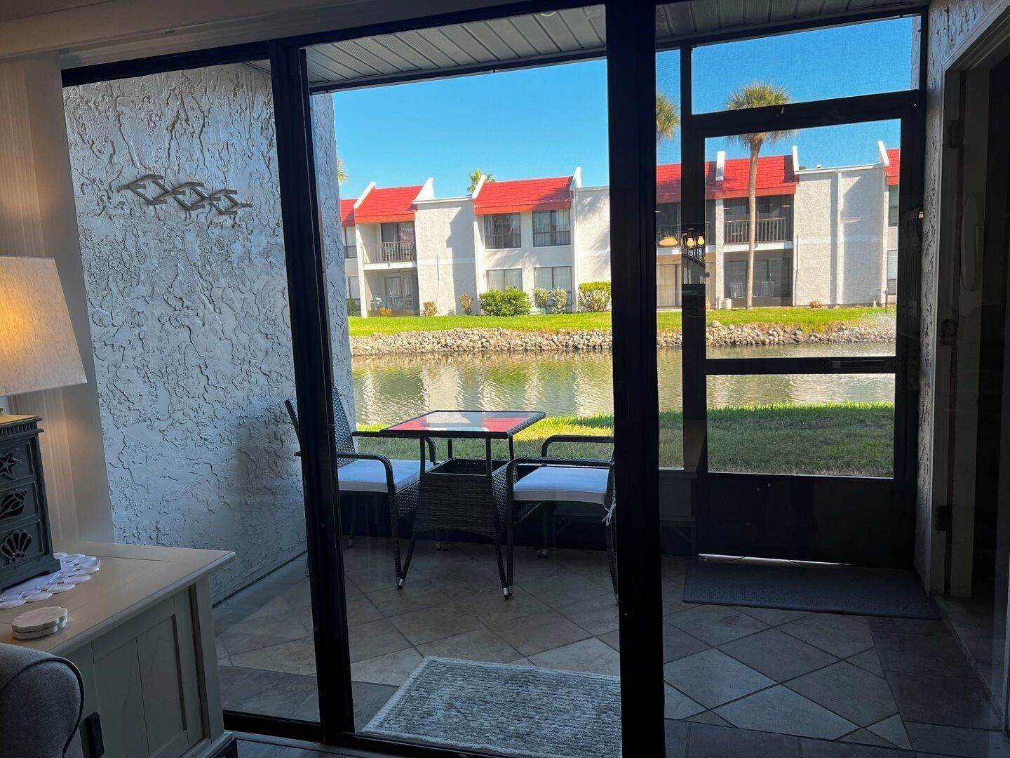 Habitación con puerta de vidrio que da a un patio exterior con mesa y sillas, vista a un edificio de tejados rojos y una masa de agua.